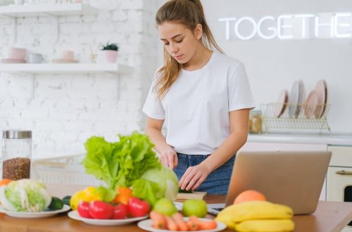 Woman in white t-shirt preparing fresh vegetables in a bright kitchen with a laptop.