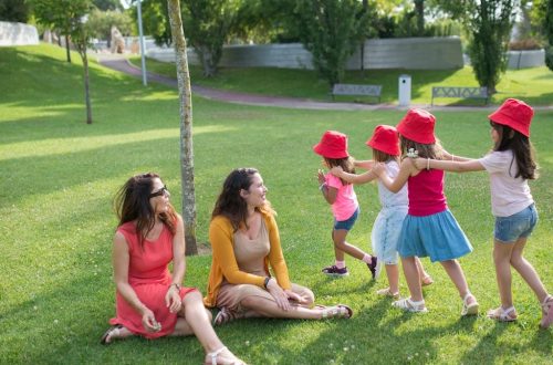 Happy children playing in a sunny park with two mothers sitting nearby, enjoying the outdoors.